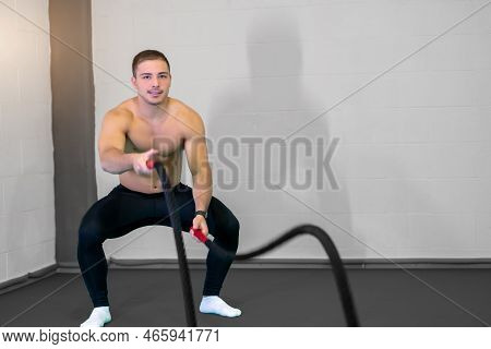 A Handsome Muscular Young Man Using Battle Ropes For Exercise In A Gym. Look At Camera. Motivational