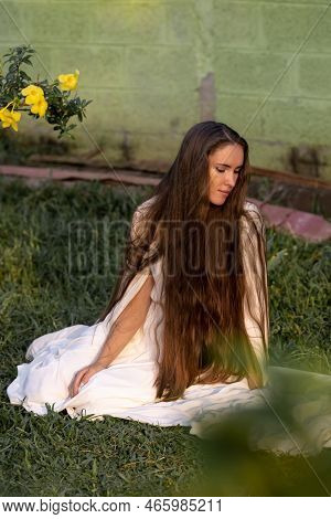 A Girl With Very Long Hair Sits On The Grass In A Torpic Landscape. White Dress Of The Bride. Health