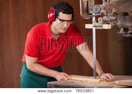 Young male carpenter cutting wooden plank with bandsaw in workshop