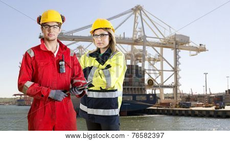 Two dockers, wearing safety gear, protection and work clothing, posing in front of a large container ship in an industrial harbor