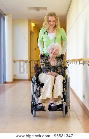 Nurse Assisting Elderly On Wheelchair At Corridor
