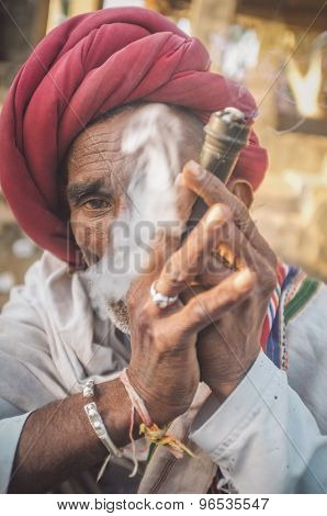 GODWAR REGION, INDIA - 14 FEBRUARY 2015: Elderly Rabari tribesman with red turban and blanket smokes chillum. Post-processed with grain, texture and colour effect.