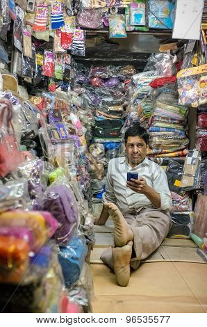 JODHPUR, INDIA - 07 FEBRUARY 2015: Store owner sitting on floor of clothing shop with mobile phone while waiting for customers. Little local textile shop overflown with different merchandise.