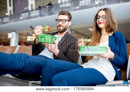 Elegant business couple eating with lunch boxes sitting at the waiting hall in the airport. Having a snack during business trip