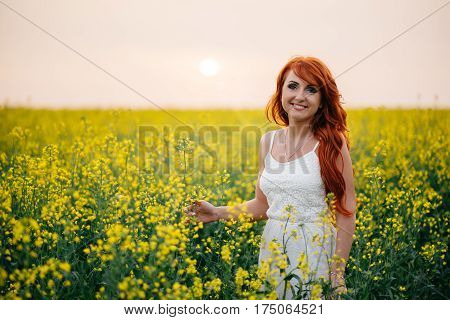 Young beautiful red-haired woman in a blooming rapeseed field at sunset