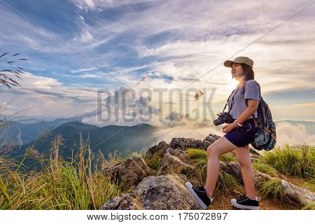 Hiker asian young woman happy with camera backpack and caps looking beautiful landscape nature of mountain and colourful sky at sunset on viewpoint Phu Chi Fa Forest Park Chiang Rai Thailand