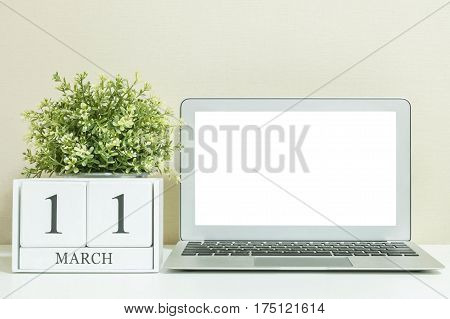 White wooden calendar with black 11 march word with white blank space at center of computer notebook on white wood desk and cream wallpaper textured background selective focus at the calendar