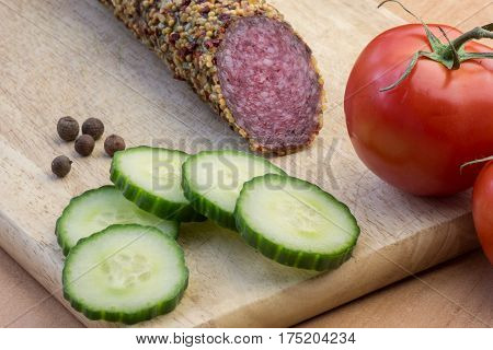 Delicious smoked sausage whole on wooden board with slices of cucumbers and tomatoes. View from above.