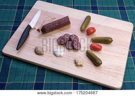 Delicious smoked sausages whole and sliced on wooden cutting board with cucumbers, tomatoes and slices of onions. View from above.
