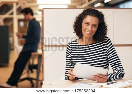 Portrait of a smiling businesswoman sitting at a desk in a modern office working on a digital tablet with a colleague working in the background