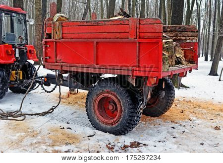Red trailer with firewood attached to the tractor in the winter in the wood or the park. Side view.