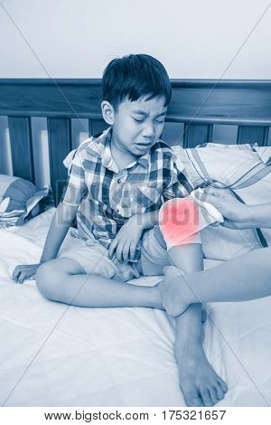Child injured. Mother bandaging son's knee on bed bandage in focus. Asian boy sadden and crying. Human health care and medicine concept. Color increase blue skin and red spot indicating of pain.