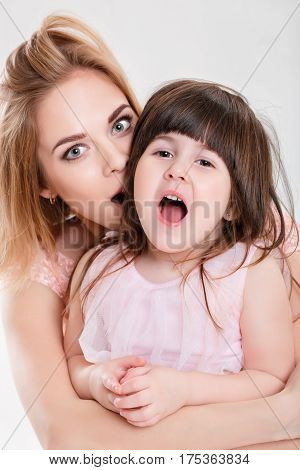 portrait of blonde mom and sweet little daughter in pink dresses princesses hugging and laughing on gray background. family
