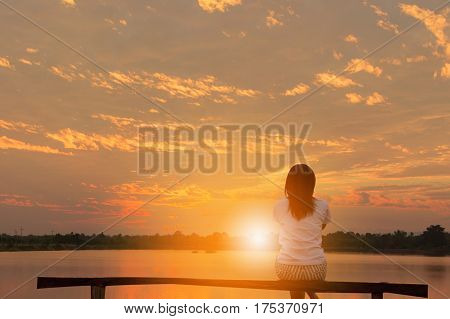 Beautiful woman sitting relaxed on a wooden looking sunset at a lake background.