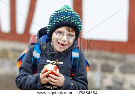 Little kid boy with eye glasses on the way to the school and eating fresh apple. Child with missing front teeth wearing warm clothes, glasses and backpack. Happy student or people. Education concept