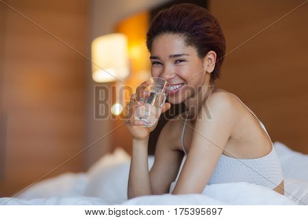 Young beautiful woman with glass of water in a bed. Smiling woman laying in the hotel room. Young happy black woman sits in a bed in the hotel.