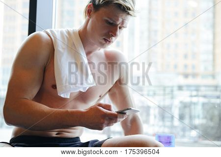 Atletic man with naked torso which sitting near the window and using smartphone