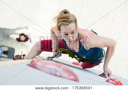 Cute female Athlete hanging on climbing Wall making hard move and belaying Partner watching from below. National Climbing competitions, Dnipro, Ukraine, May 20, 2016
