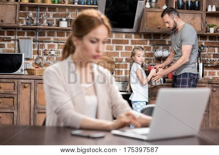 Young father and child girk making tea while woman working on laptop