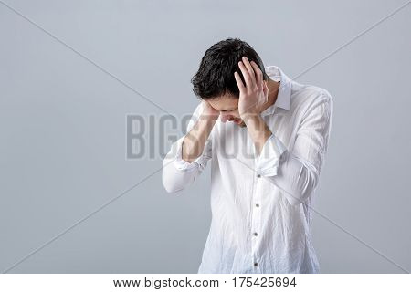 Frustrated young brunette man in a white shirt with headache on  gray background. terrible migraine