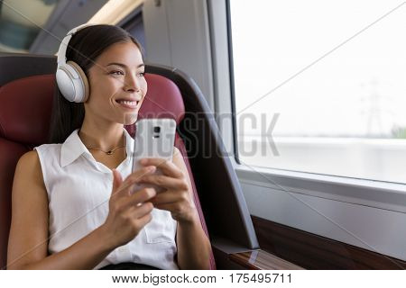 Woman on train listening to music on smartphone. People lifestyle. Young urban businesswoman using phone app, wireless headphones to listen to audiobook. Asian girl enjoying travel in business class.