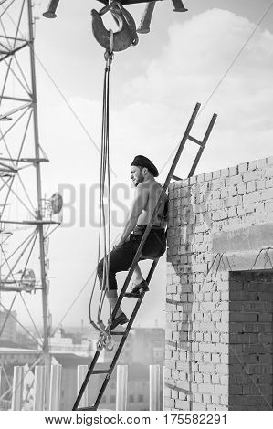 Vintage construction. Vertical monochrome shot of a muscular shirtless vintage handy man sitting relaxing on a ladder at construction site looking away rearview building industry business confident