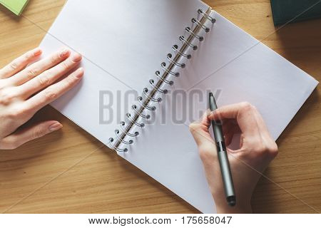 Top view of white girl's hand writing in empty spiral notepad placed on wooden desktop. Copy space