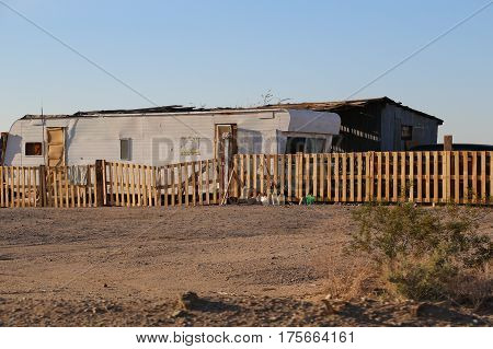 Delapidated mobile home and shed behind fence in rural desert, probablyowned by family of low income, in poverty