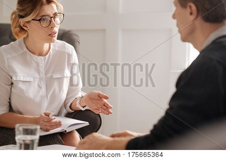 Psychological session. Professional confident intelligent therapist holding a notebook and speaking to her patient while having a psychological session with him