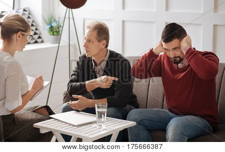I do not want to hear you. Serious stubborn bearded man sitting on the sofa and pressing his hands to the ears while being unwilling to listen