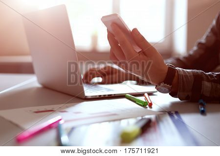 Side View of Man holding Telephone in front of Laptop in dark warm tones with focus on edge of phone. Color Markers and Pens dropped on Desk Sunlight on Background