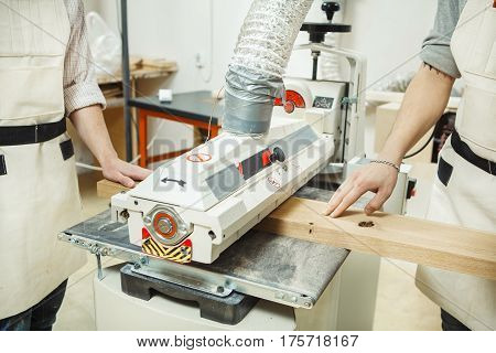 Two turners from different sides holding wooden bar on drum-grinding machine with oscillation. Machine-tool equipped with hood. Men dressed in overalls.