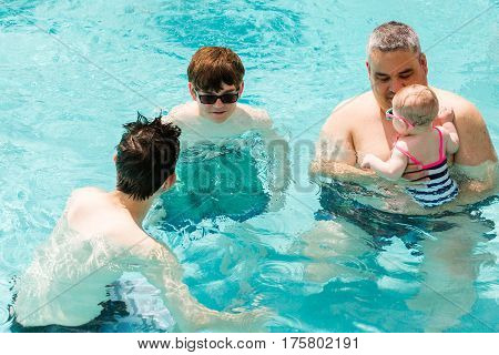 Family with cute baby girl faving fun in outdoor swimming pool on hot summer day.