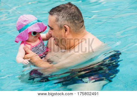 Family with cute baby girl faving fun in outdoor swimming pool on hot summer day.