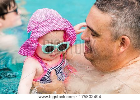 Family with cute baby girl faving fun in outdoor swimming pool on hot summer day.
