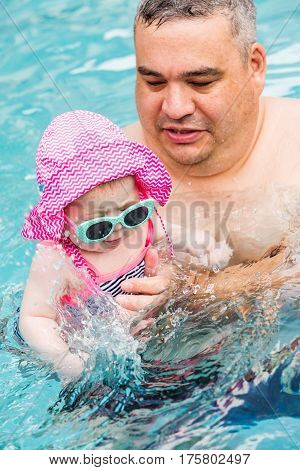 Family with cute baby girl faving fun in outdoor swimming pool on hot summer day.