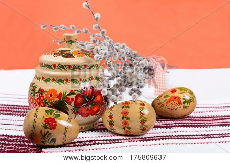 Hand painted wooden pot and Easter eggs with willow on embroidered tablecloth. Orange background. Ukrainian national painting petrikovka. Close shot.