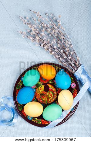 Hand painted wooden plate with colorful eggs willow with lovely polka dot ribbon on blue fabric tablecloth. Easter background. Ukrainian national painting petrikovka. Vertical orientation.