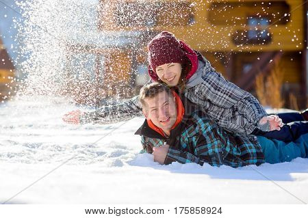 Young couple lies on the snow. The girl climbed onto the back of her boyfriend and sprinkled it with snow. Between them comic fight. Sunny winter day. Good mood. Young people smile