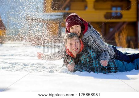 Young couple lies on the snow. The girl climbed onto the back of her boyfriend and sprinkled it with snow. Between them comic fight. Sunny winter day. Good mood. Young people smile
