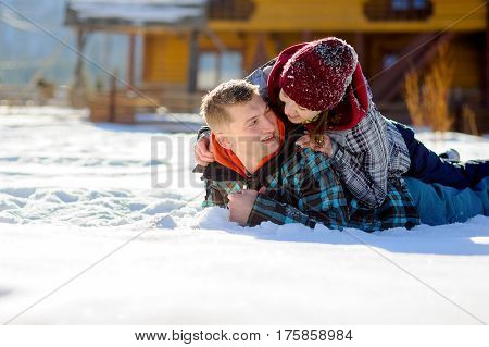 Young couple lies on the snow. Girl climbed onto the back of her boyfriend. Young people look at each other with love and tenderness. Sunny winter day. Good mood.