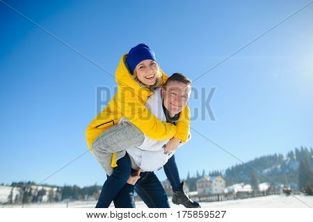 Young man holding his girlfriend on his shoulders. Girl embraces guy for neck. Young people look in the camera and cheerfully smile. Against the background of the blue sky.