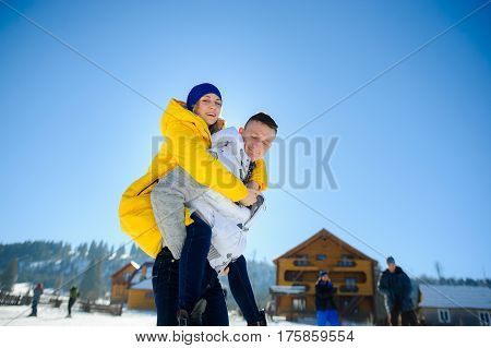 Young man holding his girlfriend on his shoulders. Girl embraces guy for neck. Young people look in the camera and cheerfully smile. Against the background of mountains and blue sky.