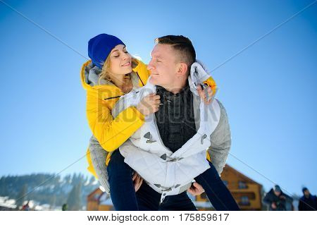 Young man holding his girlfriend on his shoulders. Girl embraces guy for neck. Young people look at each other with love and tenderness. Against the background of the blue sky.