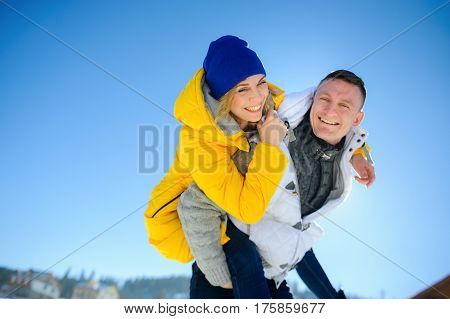 Young man holding his girlfriend on his shoulders. Girl embraces guy for neck. Young people look in the camera and cheerfully laugh. Against the background of the blue sky.