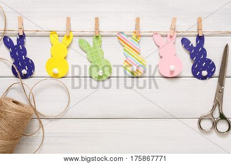 Easter bunny decoration paper cut background. DIY holiday handicraft garland of colorful rabbits and craft tools. Top view with copy space on white wood