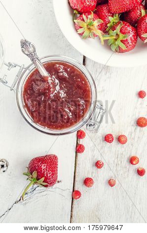 Homemade delicious strawberry jam in a glass jar and fresh strawberries on white rusric wooden background. Top view.