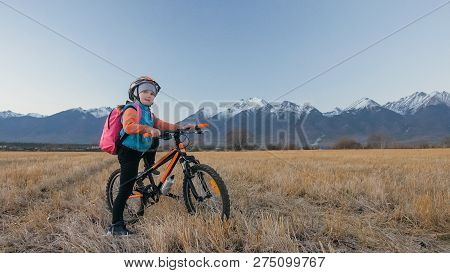 One Caucasian Children Walk With Bike In Wheat Field. Little Girl Walking Black Orange Cycle On Back