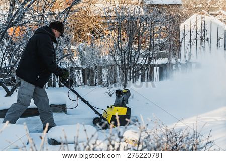 A Man In A Black Jacket And A Gray Pants Is Brushing White Snow With The Yellow Electric Snow Throwe