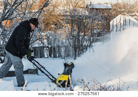 A Man In A Black Jacket And A Gray Pants Is Brushing White Snow With The Yellow Electric Snow Throwe